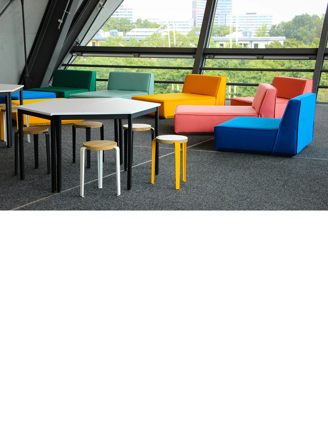 Bunte modulare Sofalandschaft und schlichte Tische mit Hockern in einem lichtdurchfluteten Meetingbereich mit Ausblick auf eine grüne Stadtlandschaft.
