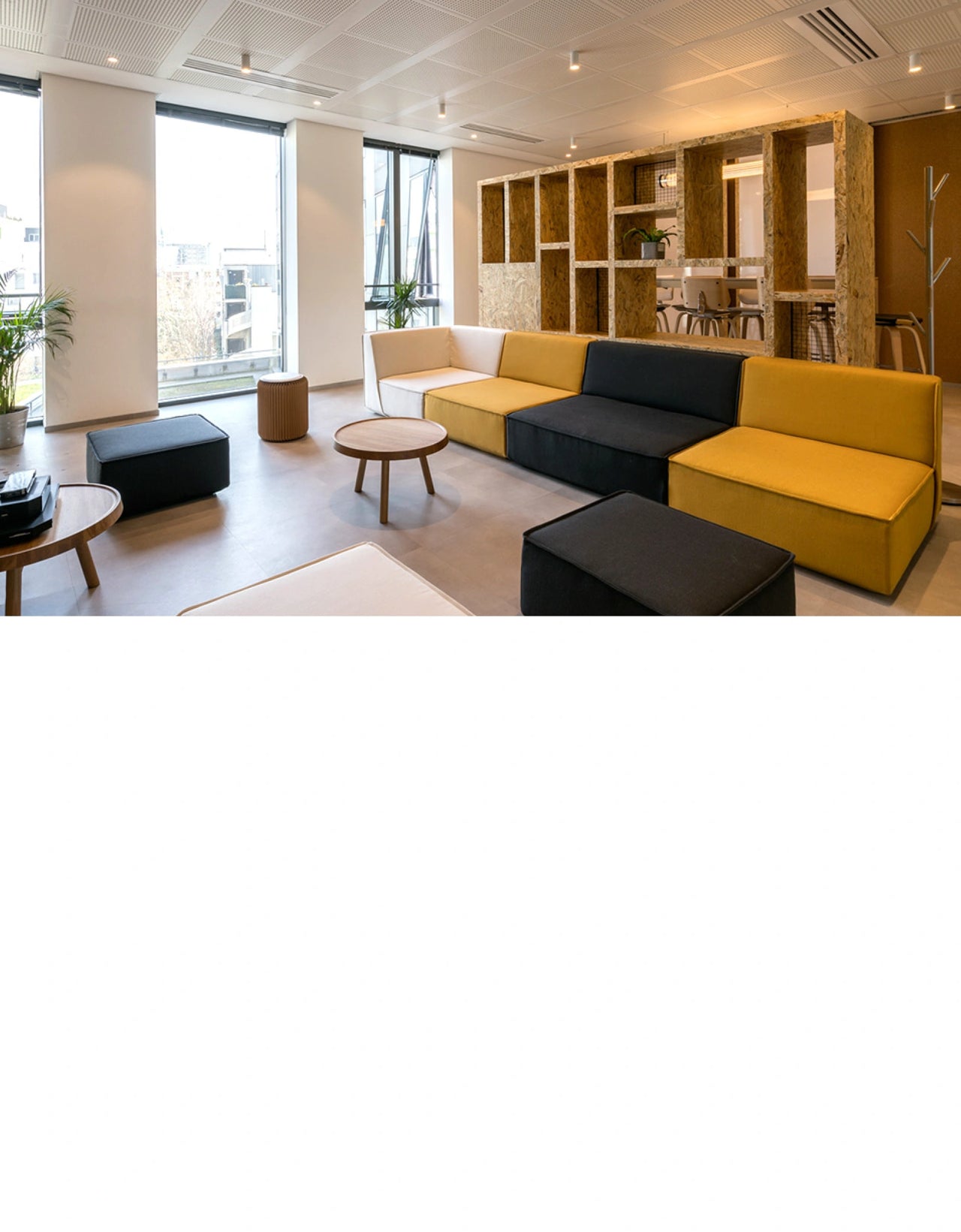 Moderne Lounge-Ecke in einem Büro mit modularen Sofaelementen in Weiß, Gelb und Schwarz, ergänzt durch runde Holztische und große Fenster mit Blick nach draußen.