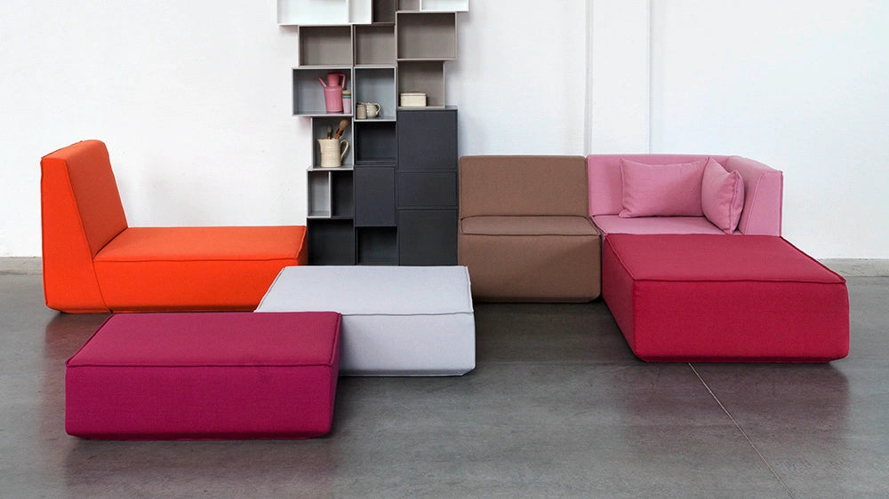A versatile Cubit sofa module combination in orange, pink, berry, and brown, arranged in front of a tall, gray Cubit Shelving system a modern industrial studio.