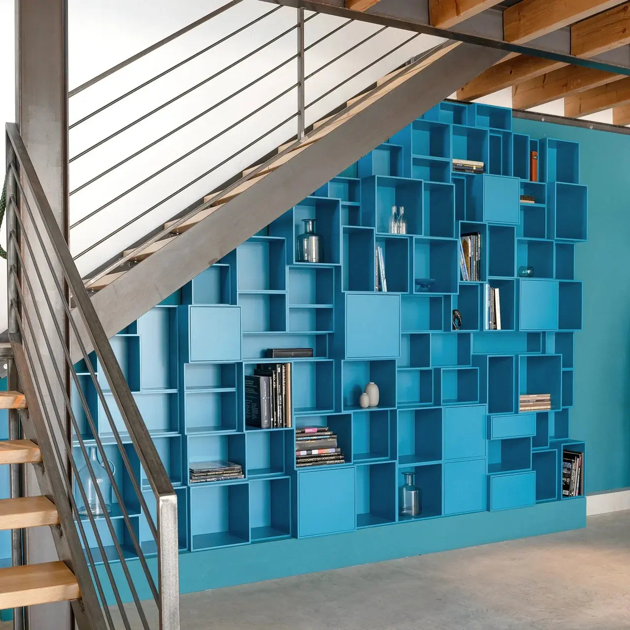 Bodentiefes modulares Regalwand-System in Petrolblau unter einer offenen Stahltreppe – individuell konfigurierbare Cubes mit Büchern und Dekobjekten in einem Loft-Interieur.