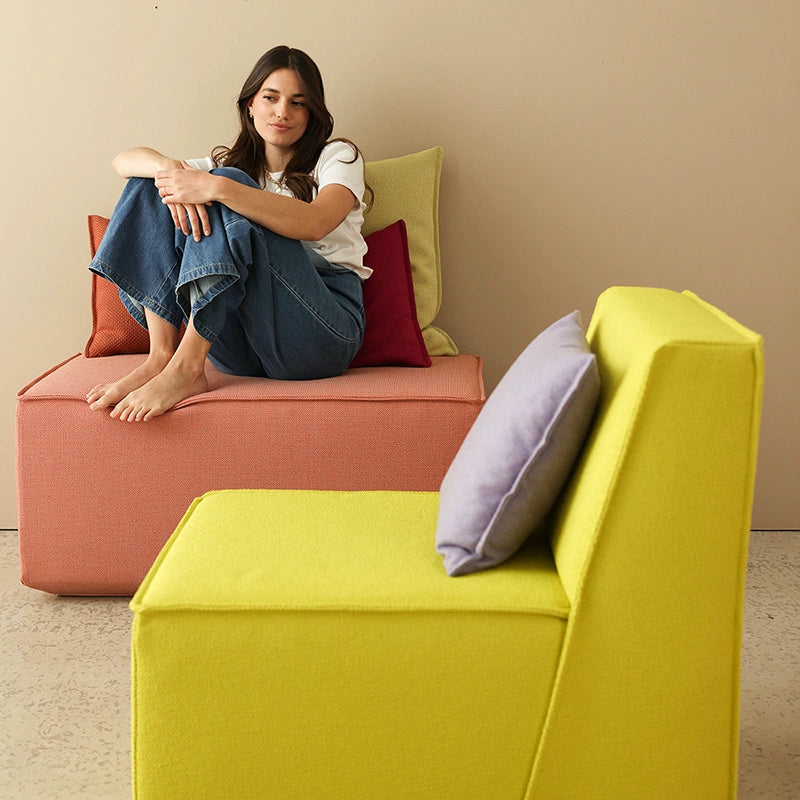 A young woman is sitting on a colorful coral-and-yellow sofa set with various throw pillows in front of a beige wall.