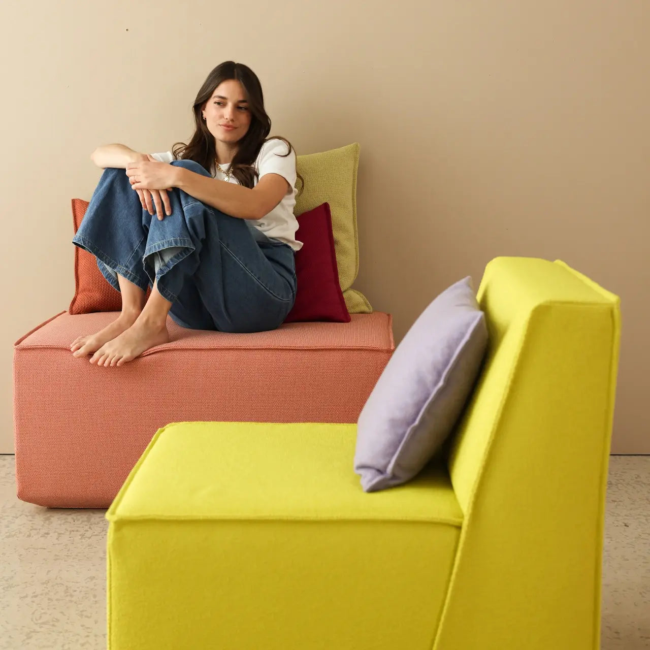 Modulares Sofa-System in Koralle und Gelbgrün mit bunten Kissen – flexibel kombinierbares Sitzmöbel für das Wohnzimmer, präsentiert von einer Frau.