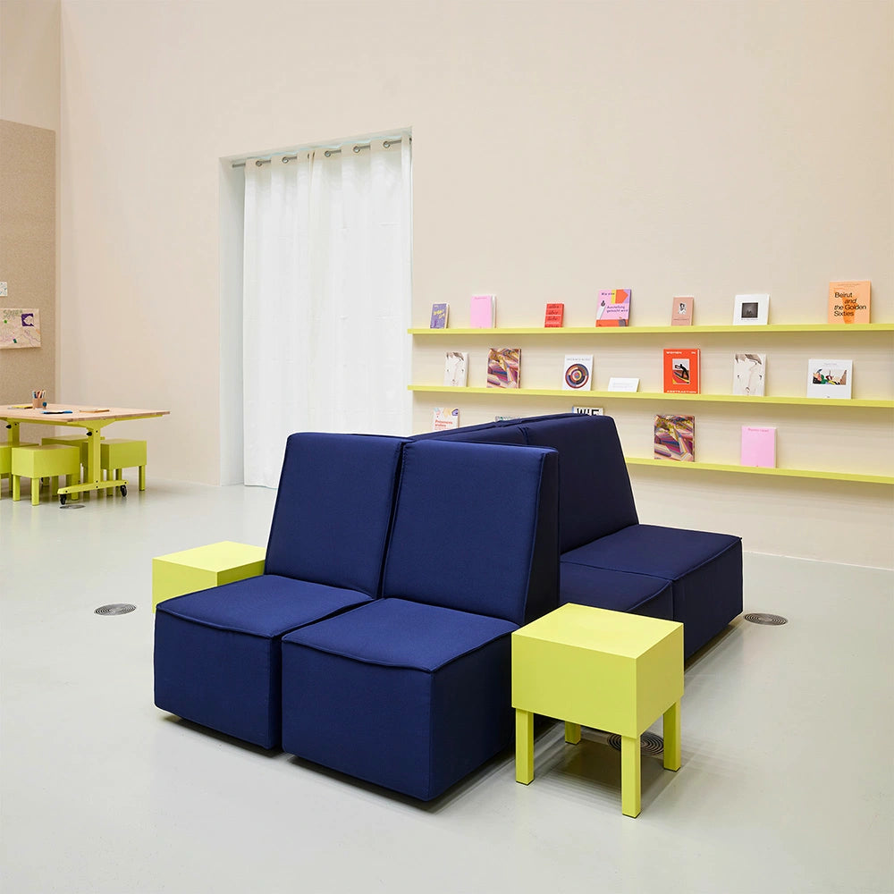 A two-sided Cubit sofa island in dark blue, paired with neon yellow side tables and stools in a minimalist showroom featuring yellow wall shelves.