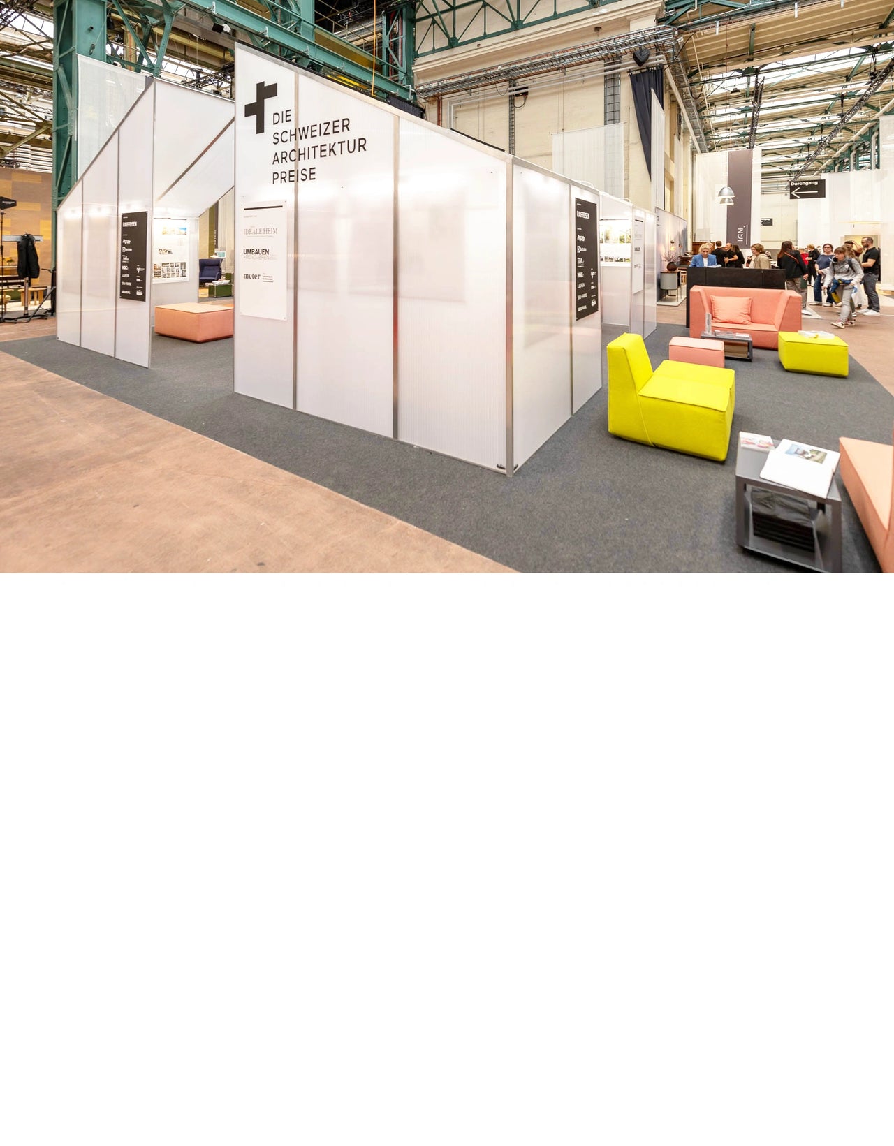 Stand des Prix suisses d'architecture dans un hall industriel, équipé de sièges modulaires jaunes, roses et orange pour créer une ambiance lounge accueillante.