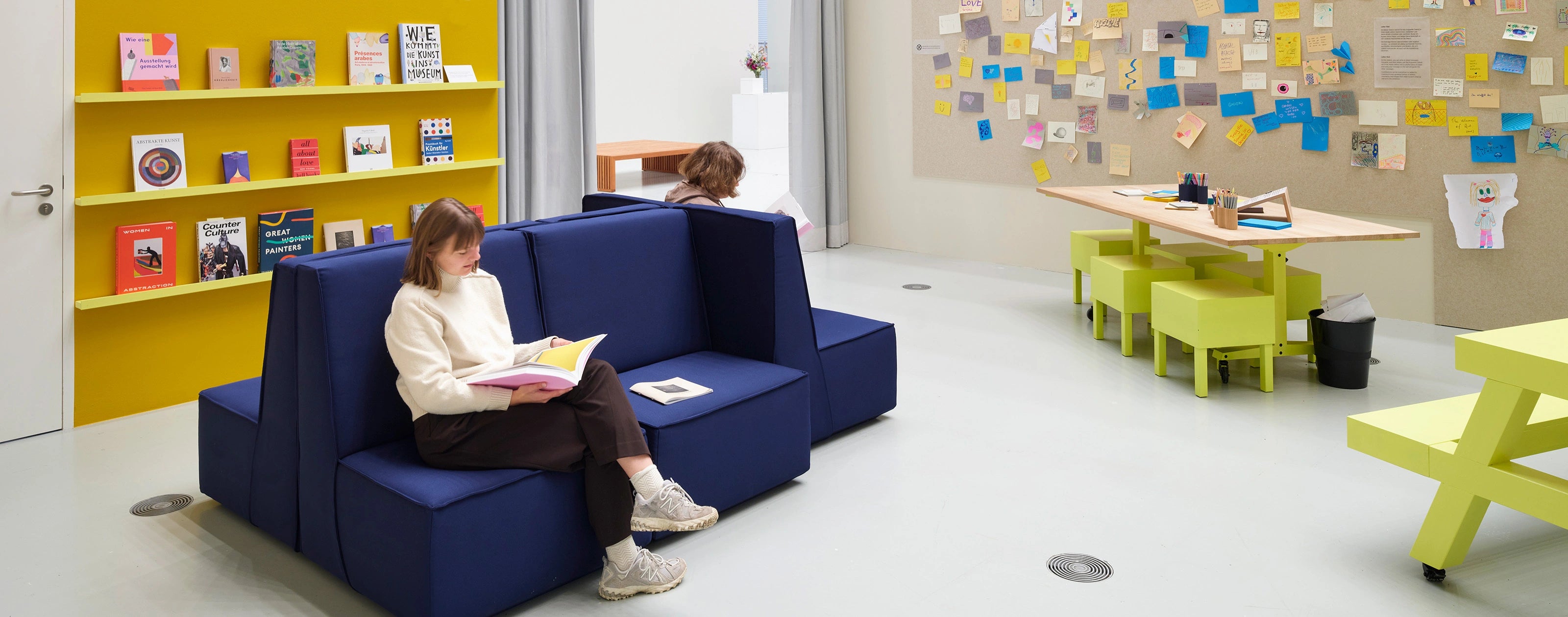 Une pièce lumineuse avec des étagères jaunes et un banc bleu sur lequel une femme lit un livre.