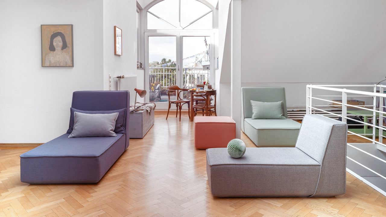 Pièce inondée de lumière du jour avec un sol en bois et différents éléments de canapés modulaires colorés en gris, bleu, vert et rouge. Au fond, une table à manger avec des chaises et une grande baie vitrée.