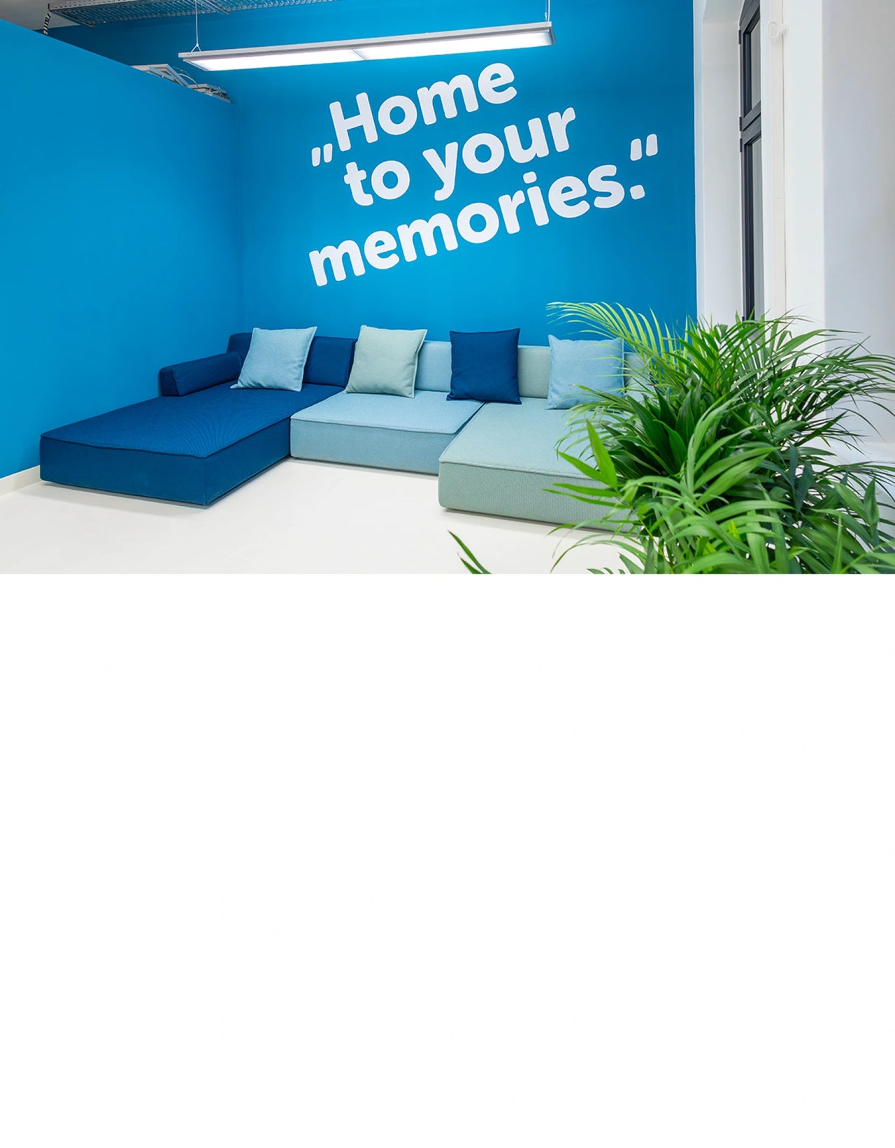 Salon moderne avec Canapé panoramique modulaire Canapé panoramique les tons bleus devant un mur bleu vif portant l'inscription « Home to your memories », complété par des plantes vertes dans la zone d'accueil.