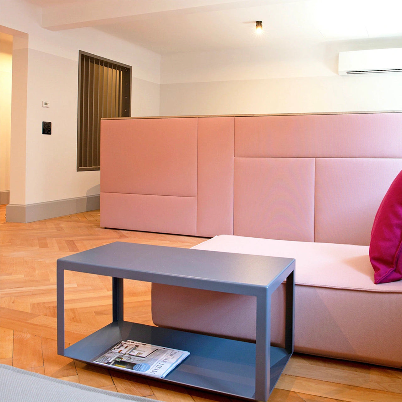 Salle d'attente moderne avec comptoir acoustique rose, banquette assortie, table basse gris foncé et parquet à chevrons.